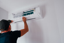 Service And Maintenance Of The Air Conditioner. A Young Male Technician Is Cleaning And Replacing The Air Conditioner Filter Against The Background Of A White Wall