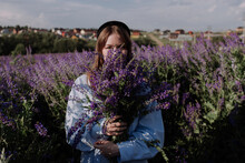 Young Woman In Shirt Holding Bouquet Of Purple Flowers Covering Face Standing Alone Among Field In Front Of Blue Sky Enjoying Of Sunlight At Golden Hour. Summer Outfit. Girl With Flowers