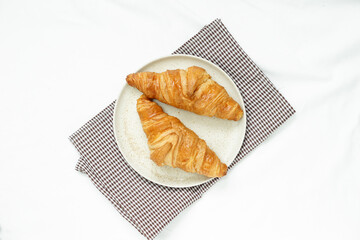 top view of  Two croissants are placed in a round ceramic  plate placed on brown checkered fabric against white crumpled tablecloths with soft light in the morning.