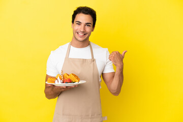 Sticker - Restaurant waiter holding waffles over isolated yellow background pointing to the side to present a product