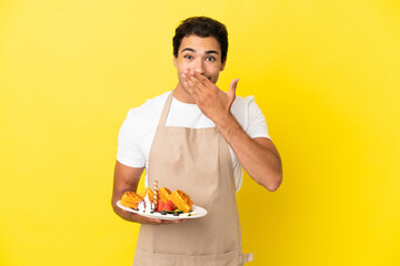 Sticker - Restaurant waiter holding waffles over isolated yellow background happy and smiling covering mouth with hand