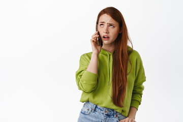 Annoyed teen girl with red hair talks on phone, roll eyes bothered and irritated by boring conversation, standing displeased against white background