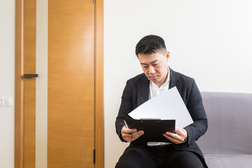 Young successful Asian man, waiting for a job interview, in the waiting room of the office center, sitting on a chair near the reception of the office center