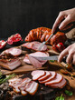 Hands cutting cold meat product, ham, sausage, salami, parma, prosciutto, bacon on wooden cutting board with herb and spices over dark background. Meat appetizer, set of wine, close up