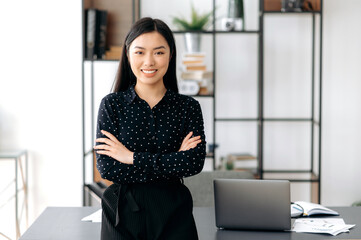 Portrait of successful confident pretty, young asian business woman or freelancer standing near work desk in office, wearing formal stylish clothes, arms crossed, looking at camera, smiling friendly