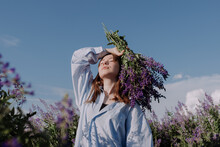 Smiling Young Woman In Shirt Holding Bouquet Of Purple Flowers Standing Alone Among Field In Front Of Blue Sky Enjoying Of Sunlight At Golden Hour. Summer Outfit. Girl With Flowers And Closed Eyes