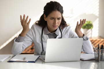 Angry female nurse work on laptop in clinic frustrated by slow internet connection on gadget. Unhappy mad woman doctor use computer in hospital confused with operational problem or spam on device.