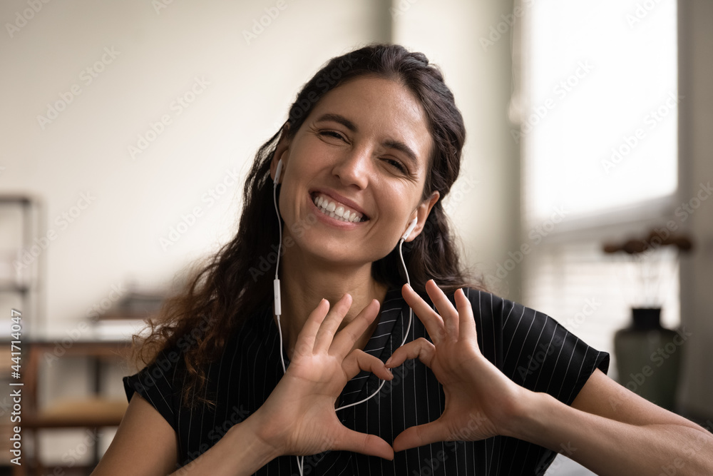 Close up headshot portrait of smiling young Caucasian female in headphones talk on video call laugh show love heart hand gesture. Happy millennial woman have webcam virtual digital communication. - obrazy, fototapety, plakaty 