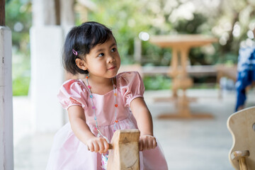 Wall Mural - Adorable Little Asian girl in pink dressed sitting on wooden horse chair.