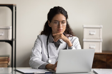 Poster - Young Caucasian female doctor in white medical uniform look at laptop screen work online in modern private hospital. Pensive woman GP use computer in clinic consult patient distant on gadget thinking.