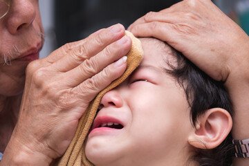 Close up of a 1-2 year old Asian face boy is crying from pain in his forehead. Grandfather uses ice wrapped in a cloth to cool the injured area. Elementary Nurse Practitioner.