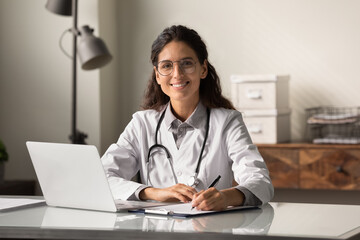 Poster - Portrait of smiling young Caucasian female doctor or nurse in white medical uniform in clinic fill journal. Happy woman GP work online consult patient on computer in hospital. Healthcare concept.