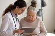 © fizkes - Young Caucasian female doctor and old 60s woman patient look at tablet screen discuss prescription together. GP or therapist consult mature client in private clinic using modern pad device.