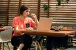 © toxicoz - Portrait of confident mature professional woman in glasses, a coral T-shirt sitting on summer terrace in cafe, using laptop computer for work, laughing happily indoors