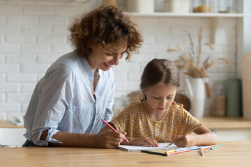 Happy young mommy drawing pictures in paper album with adorable small kid daughter, sitting at table in morn kitchen. Joyful family involved in art creative activity, early children development.