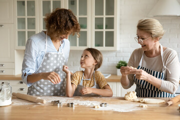 Wall Mural - Happy young 35s woman talking enjoying cooking homemade pastry with joyful cute little preschool kid daughter and caring beautiful older retired mother in modern kitchen, family culinary activity.