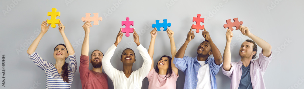 Group of six happy smiling young people holding big colorful jigsaw puzzle pieces standing against light grey studio background. Team of creative millennials cooperate and find good solution together - obrazy, fototapety, plakaty 