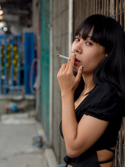 Portrait young Asian woman with long black hair, aged 20-25, wearing black shirt blue jeans. He held cigarette in his hand and smoked cigarette along the side of corridor. located outside building