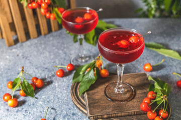 Wall Mural - Champagne coupe glass of refreshing cherry or merry cocktail with ice served on gray table surface surround of fresh merry and different green plants, shallow depth of the field.