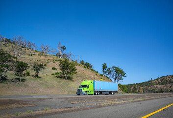 Bright lime big rig semi truck transporting cargo in dry van semi trailer moving on the summer California interstate road with die trees and dry grass on the hills