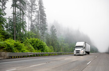 Industrial Standard Big Rig White Semi Truck Transporting Cargo In Dry Van Semi Trailer Running On The Wide Highway Road With Fog And Trees.