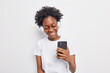 © WHstudio Leushin N - Positive teenage girl with Afro hair listens favorite song from playlist holds modern smartphone uses wireless headphones dressed in casual t shirt isolated over white background. Lifestyle leisure