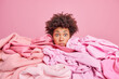 Shocked young African American woman sticks head through pile of laundry stares stunned at camera busy doing housework collects clothes for washing isolated over pink background. Clothing concept