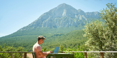 Panoramic image businessman using laptop computer keyboarding text while working remote, enjoying nature landscape with mountain view
