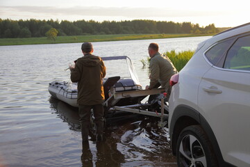 Wall Mural - Two European man unloads an inflatable motor boat from a boat trailer into the water before fishing recreation, resting on the Russian river at summer morning