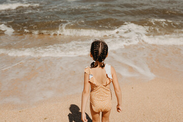 Six year girl in yellow swimsuit standing on the beach, people from behind. hot summer day.