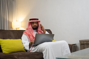 Wall Mural - an Arab businessman wearing a turban uses a cellphone to make calls and uses a laptop to work while sitting on the sofa in the office