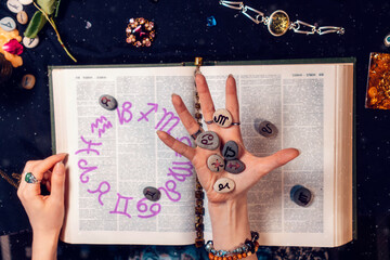 Sticker - Astrology and horoscope. The fortune teller's hand holds the zodiac stones in the palm of her hand over the open book. Top view, close-up. The concept of divination and magic