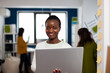 © DC Studio - Close up of african woman looking at camera smiling standing in start up creative agency office holding laptop, typing on it. Black videographer editing video project using post production software