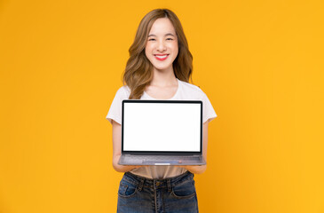 Wall Mural - Studio shot of cheerful beautiful Asian woman in white t-shirt and holding laptop mockup of blank screen on yellow background.