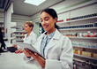 © StratfordProductions - Happy woman working in medical store wearing labcoat using digital tablet with colleague working in background