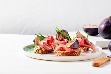 Raw ham and fresh fig with rosmarin on toasted bread on light background.