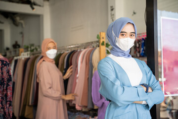 a veiled business woman wearing a mask with her arms crossed while standing in front of a boutique shop with a background of buyers choosing clothes