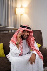 Wall Mural - Arab businessman in a turban laughing while using a cellphone while sitting on the sofa with a floor lamp and curtains as a background