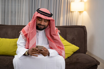 Wall Mural - Arab businessman in a turban smiling while using a cellphone while sitting on the sofa with a floor lamp and curtains as a background