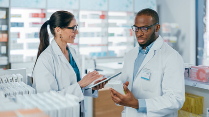 Wall Mural - Pharmacy: Black Male and Caucasian Female Pharmacists Use Digital Tablet Computer Talk about Medicine, Drugs, Vitamins, Supplements, Vaccine, Health Care Products. Medical Professionals in Drugstore