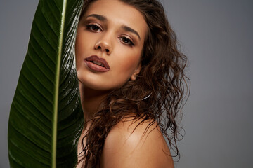 Wall Mural - Close up of beautiful naked female model looking at camera. Crop portrait of young brunette woman with perfect makeup posing with mouth open, holding big green leaf, isolated on gray.