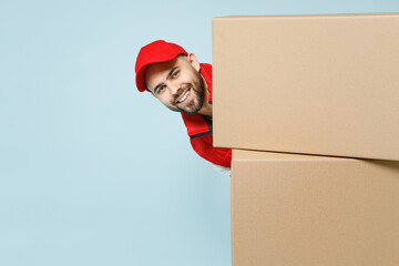 Wall Mural - Funny delivery guy employee man in red cap white T-shirt vest uniform work as dealer courier peek out from behind blank cardboard box isolated on pastel blue color background studio. Service concept.