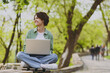 © ViDi Studio - Full length young fun happy student freelancer woman 20s in green jacket jeans sit on bench in spring park outdoors rest use laptop pc computer work online look aside People urban lifestyle concept.