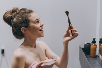 Side view amazed young excited woman 20s wear pink bra doing make up look at powder brush stand in bathroom near mirror do morning routine bodycare Skin care healthcare cosmetic procedures concept