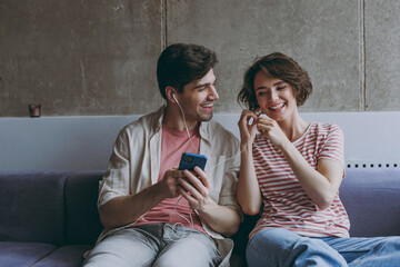 Young smiling couple two friends woman man in casual clothes earphones sitting on sofa use mobile cell phone listen music new playlist resting indoors at home flat together. People lifestyle concept