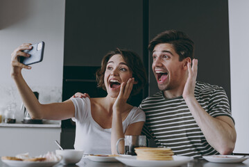 Young surprised excited couple two woman man 20s in casual clothes sit by table eat pancake do selfie by mobile cell phone hold face cook food in light kitchen at home together Healthy diet concept.