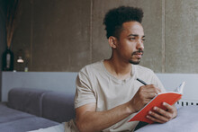 Pensive Thoughtful Young Poet Student African American Man 20s Wearing Beige T-shirt Sitting On Grey Sofa Indoors Apartment Write Down Memories In Notebook Diary, Resting On Weekends Staying At Home