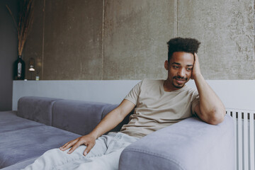 Sticker - Pensive thoughtful melancholic young african american man in beige t-shirt sweatpants sit on grey sofa indoors apartment near heating battery procrastinate prop up face, rest on weekends stay at home