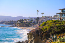 A Gorgeous Shot Of The Vast Deep Blue Ocean Water With Waves Rolling Into The Beach, Homes On The Cliffs With Lush Green Palm Trees And Plants With Blue Sky At Treasure Island Beach In Laguna Beach CA