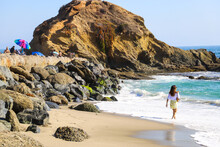 A Woman Walking On The Beach And People Relaxing Near The Rocks With Colorful Umbrellas With Vast Blue Ocean Water And Waves Crashing Into The Rocks  At Treasure Island Beach In Laguna Beach CA